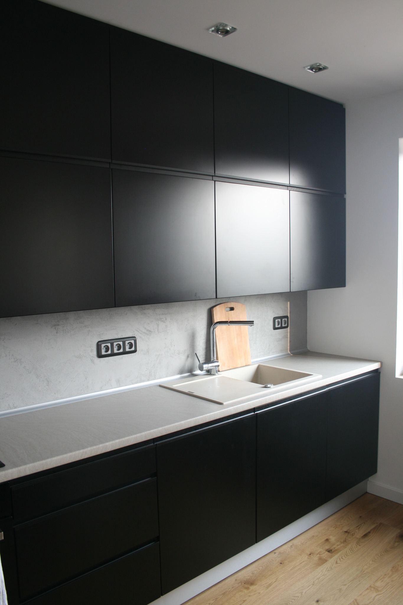 Nowoczesna kuchnia z czarnymi, matowymi szafkami, blatem w odcieniach szarości, beżowym zlewem i drewnianą deską do krojenia przy baterii.