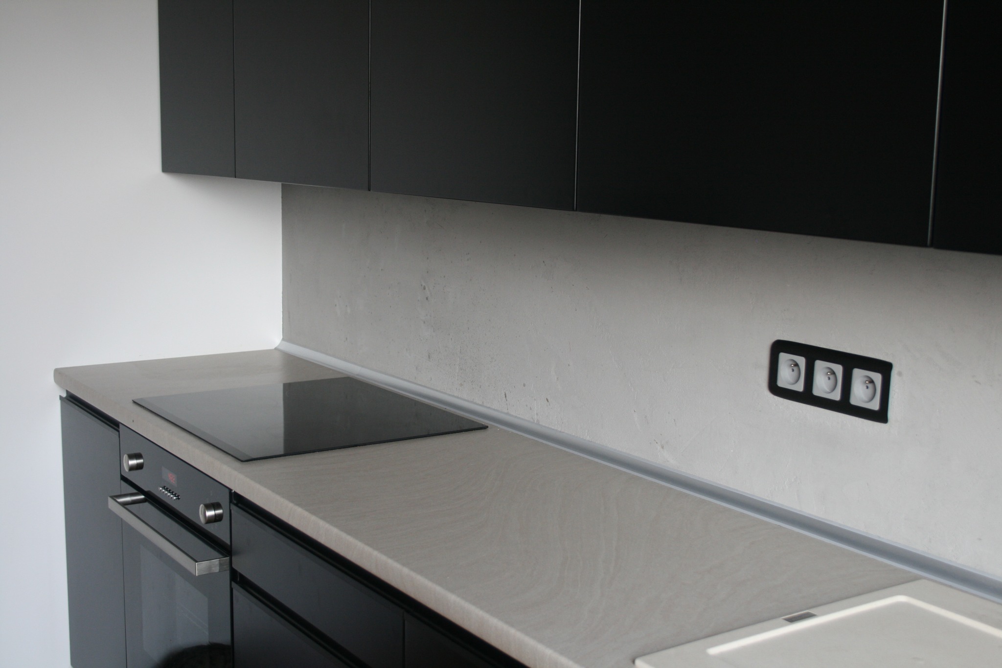 Nowoczesna kuchnia z czarnymi szafkami, szarym blatem i płytą indukcyjną. Na ścianie widoczne gniazdka elektryczne w czarnej ramce.