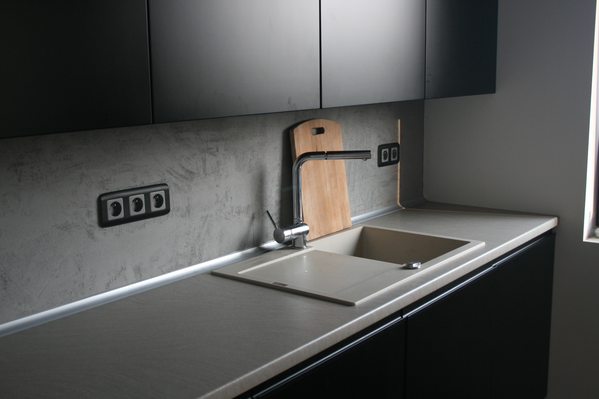 Nowoczesna kuchnia z szarymi szafkami, blatem w odcieniach beżu i zlewozmywakiem z baterią. Na ścianie widoczna deska do krojenia oraz gniazdka elektryczne w czarnych ramkach.