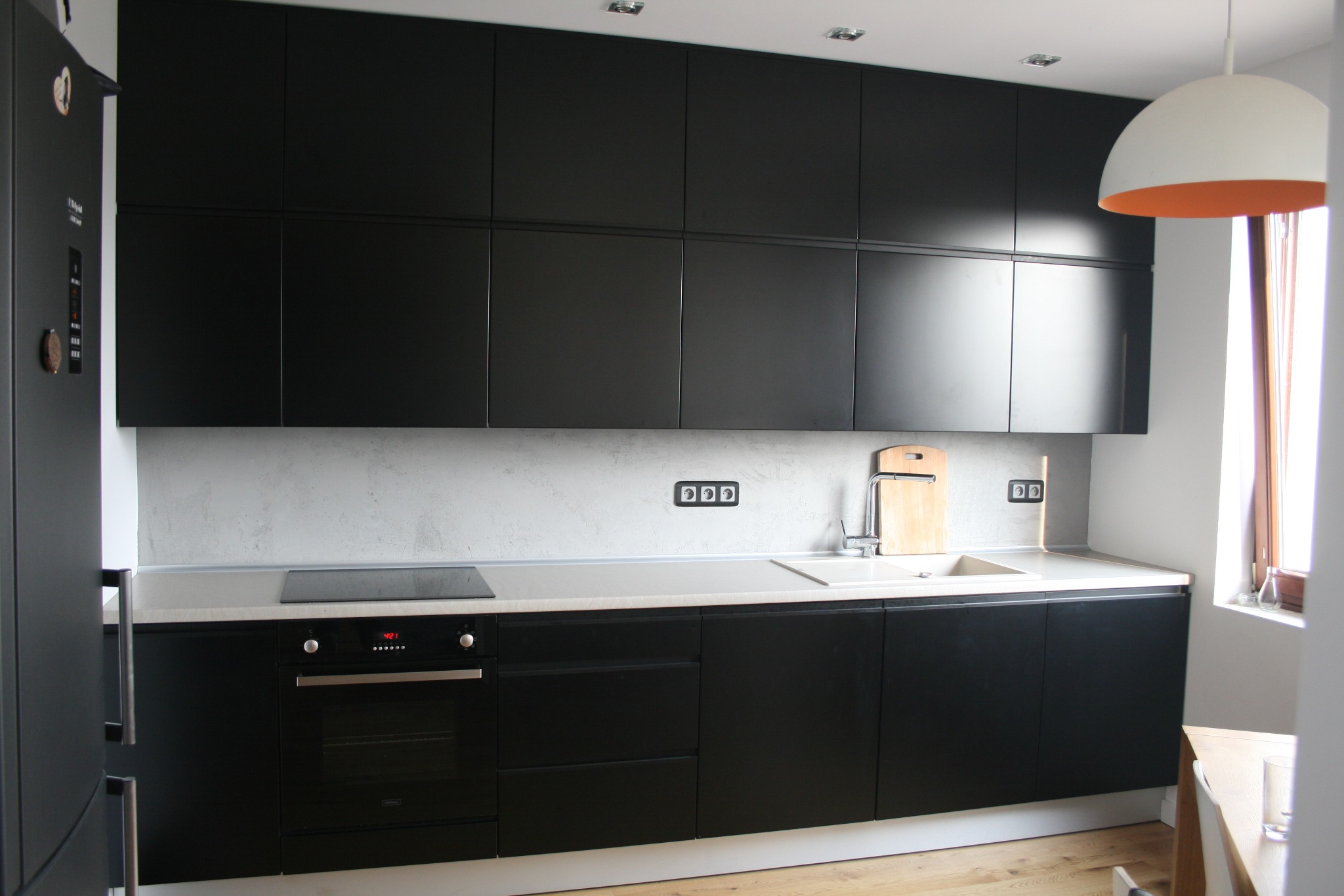 Nowoczesna kuchnia z zabudową w kolorze matowej czerni, jasnoszarym blatem i betonową ścianą nad blatem, widoczne gniazdka elektryczne w czarnej oprawie, drewniana deska do krojenia oparta o ścianę.