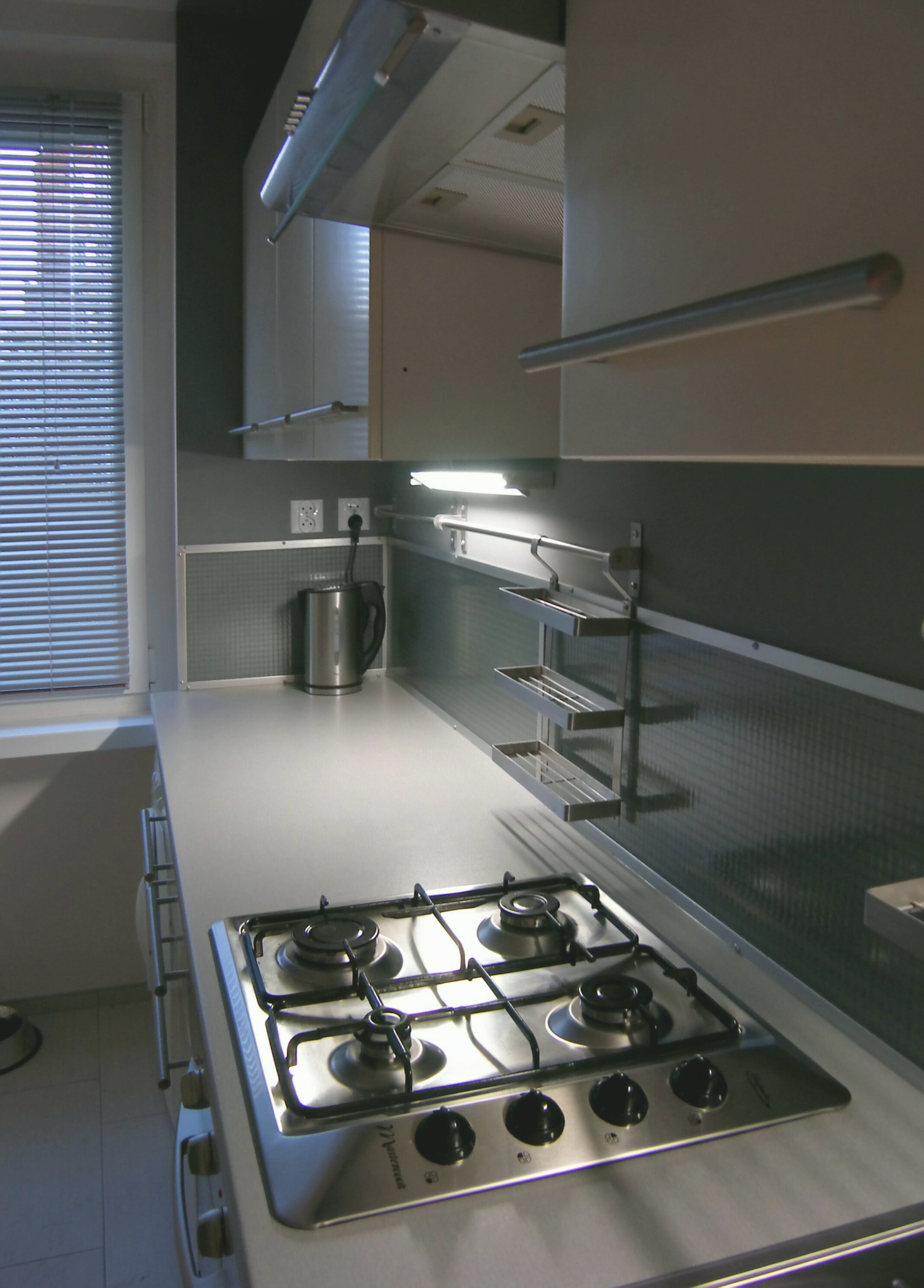 Nowoczesna kuchnia z białymi szafkami, srebrnym blatem, czteropalnikową kuchenką gazową i metalowym czajnikiem na tle okna z żaluzjami.