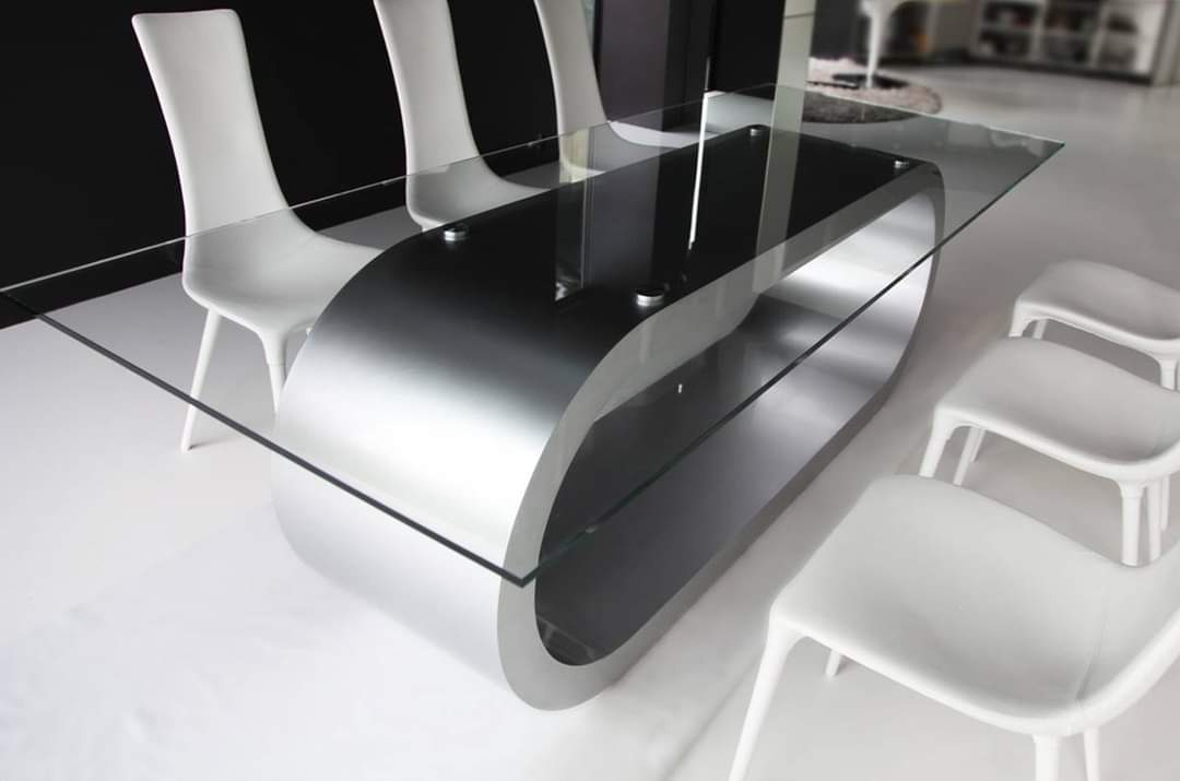 Nowoczesny stół z blatem ze szkła i metalową podstawą w kształcie wydłużonego owalu, otoczony białymi krzesłami w minimalistycznym stylu.