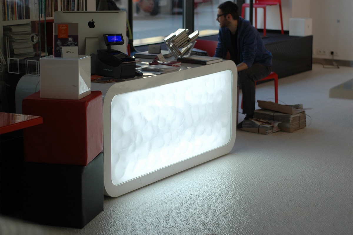 Nowoczesna lada recepcyjna z podświetlanym frontem o nieregularnej fakturze, z komputerem iMac, drukarką fiskalną i dekoracyjnym układem książek na blacie, w tle siedzący mężczyzna na czerwonym...