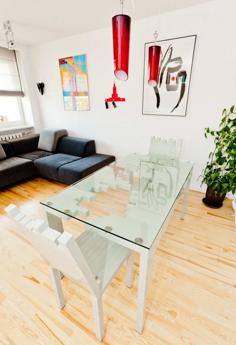 Jasne wnętrze mieszkania z drewnianą podłogą, narożną sofą i stołem z blatem z przezroczystego szkła, którego krawędzie mają dekoracyjny wzór przypominający piksele. Nad stołem wiszą dwie czerwone...