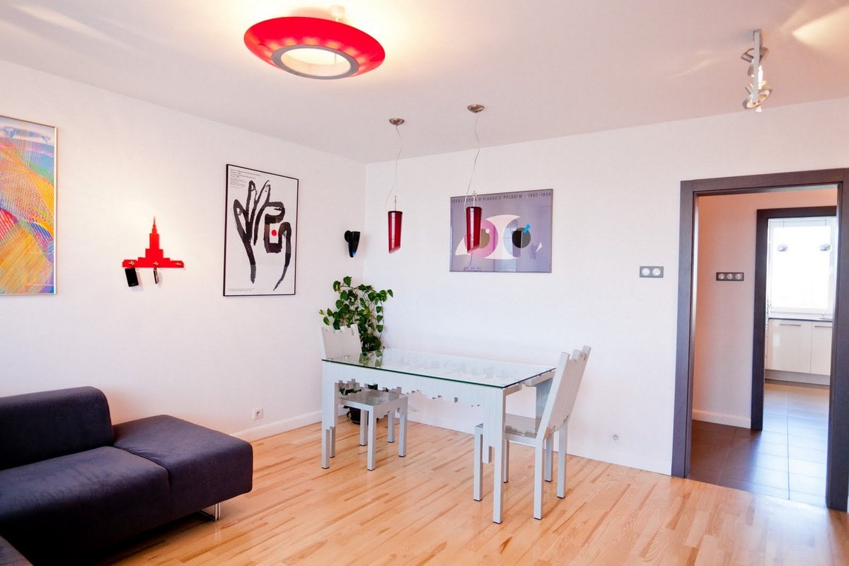 Nowoczesne wnętrze mieszkania z drewnianą podłogą, szarą sofą, białym stołem z szklanym blatem i krzesłami o ażurowej konstrukcji. Na ścianach wiszą grafiki i designerskie lampy, a w tle widoczny...