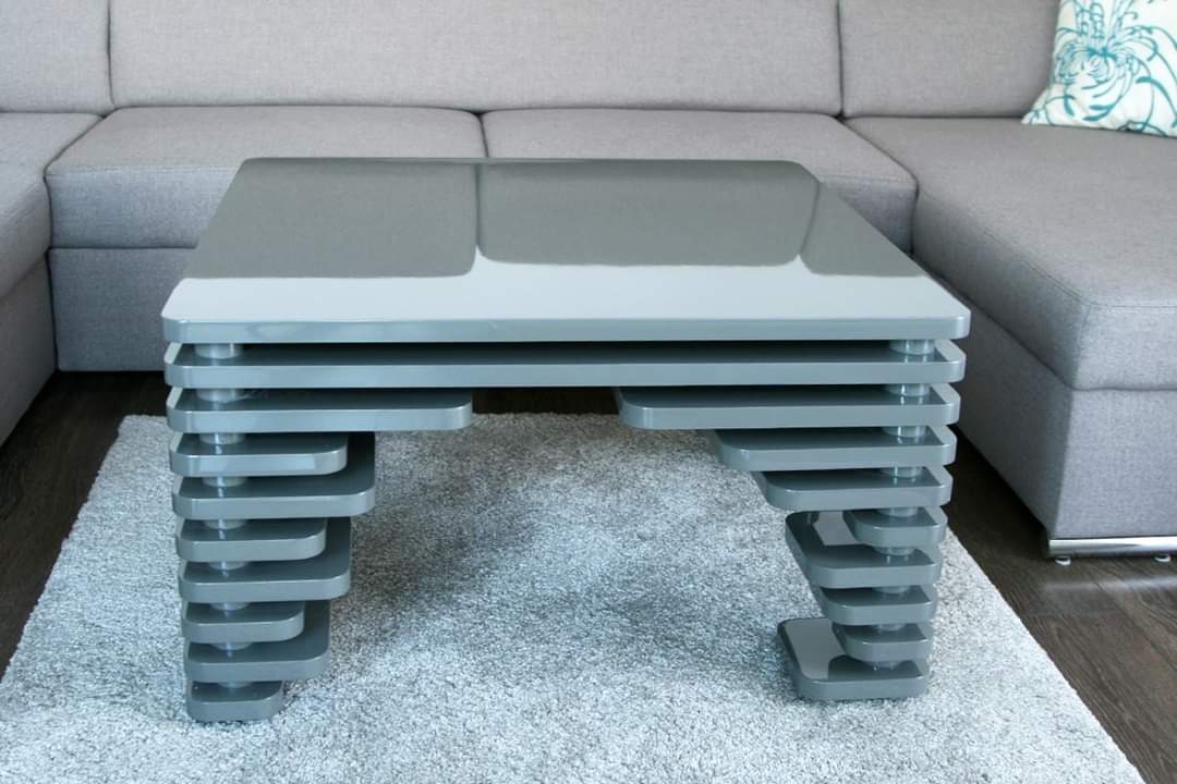 Nowoczesny, szary stolik kawowy z blatem z połyskiem i nogami składającymi się z warstwowych elementów, ustawiony na puszystym dywanie w salonie z szarą sofą.