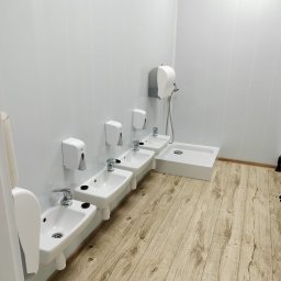 Cart Pol Sp. z o. o. - Wnętrze łazienki publicznej z czterema umywalkami i dozownikami mydła w rzędzie, obok kabina prysznicowa, ściany wyłożone płytami, podłoga imitująca drewno.