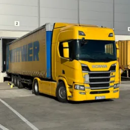 Żółta ciężarówka Scania z niebieskim napisem WITWER na naczepie, zaparkowana przed magazynem z numerem 2, z widoczną tablicą rejestracyjną DJA 35008.