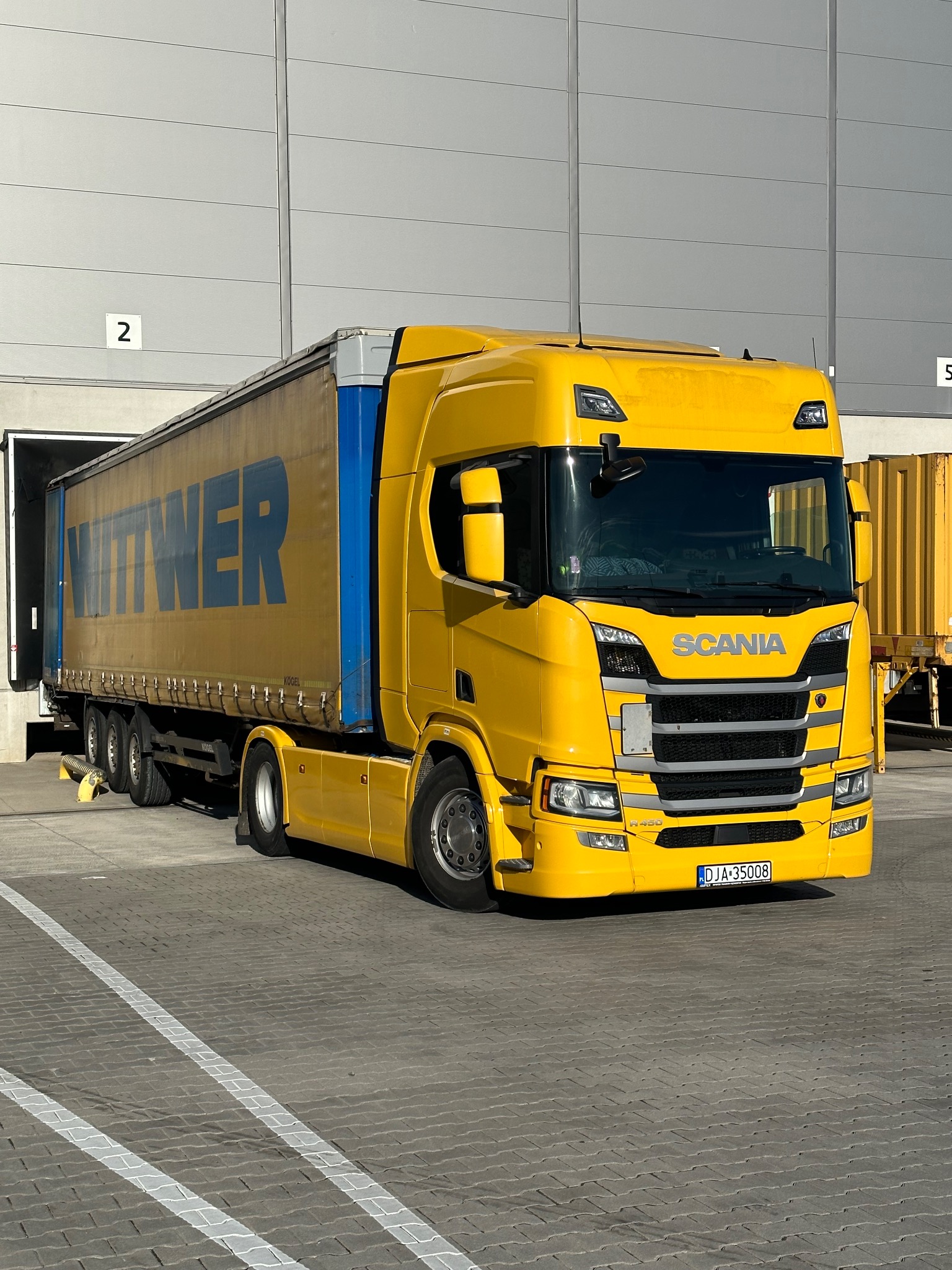 Żółta ciężarówka Scania z niebieskim napisem WITWER na naczepie, zaparkowana przed magazynem z numerem 2, z widoczną tablicą rejestracyjną DJA 35008.