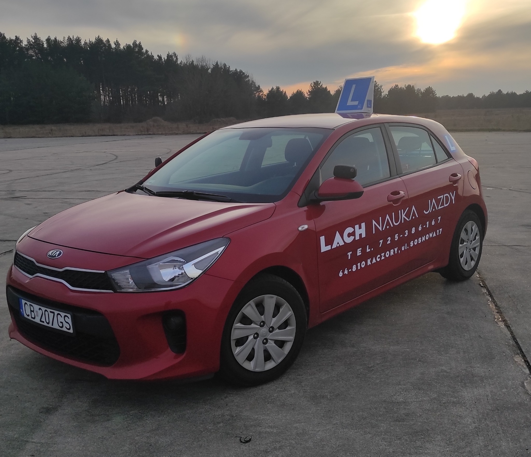 Czerwony samochód z literą 'L' na dachu, zaparkowany na betonowym placu, w tle las i zachodzące słońce. Na karoserii widnieje napis 'LACH NAUKA JAZDY' i numer telefonu.