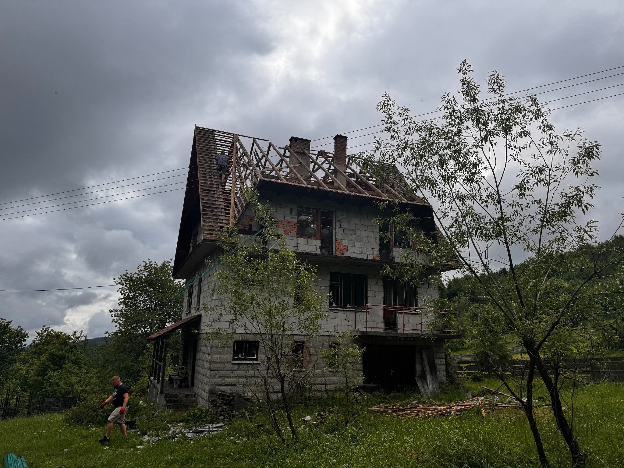 Dwupiętrowy dom w trakcie demontażu starego poszycia dachowego, widoczna więźba, murarz na dachu, ponure niebo, mężczyzna na trawniku, elementy starego dachu na trawie.