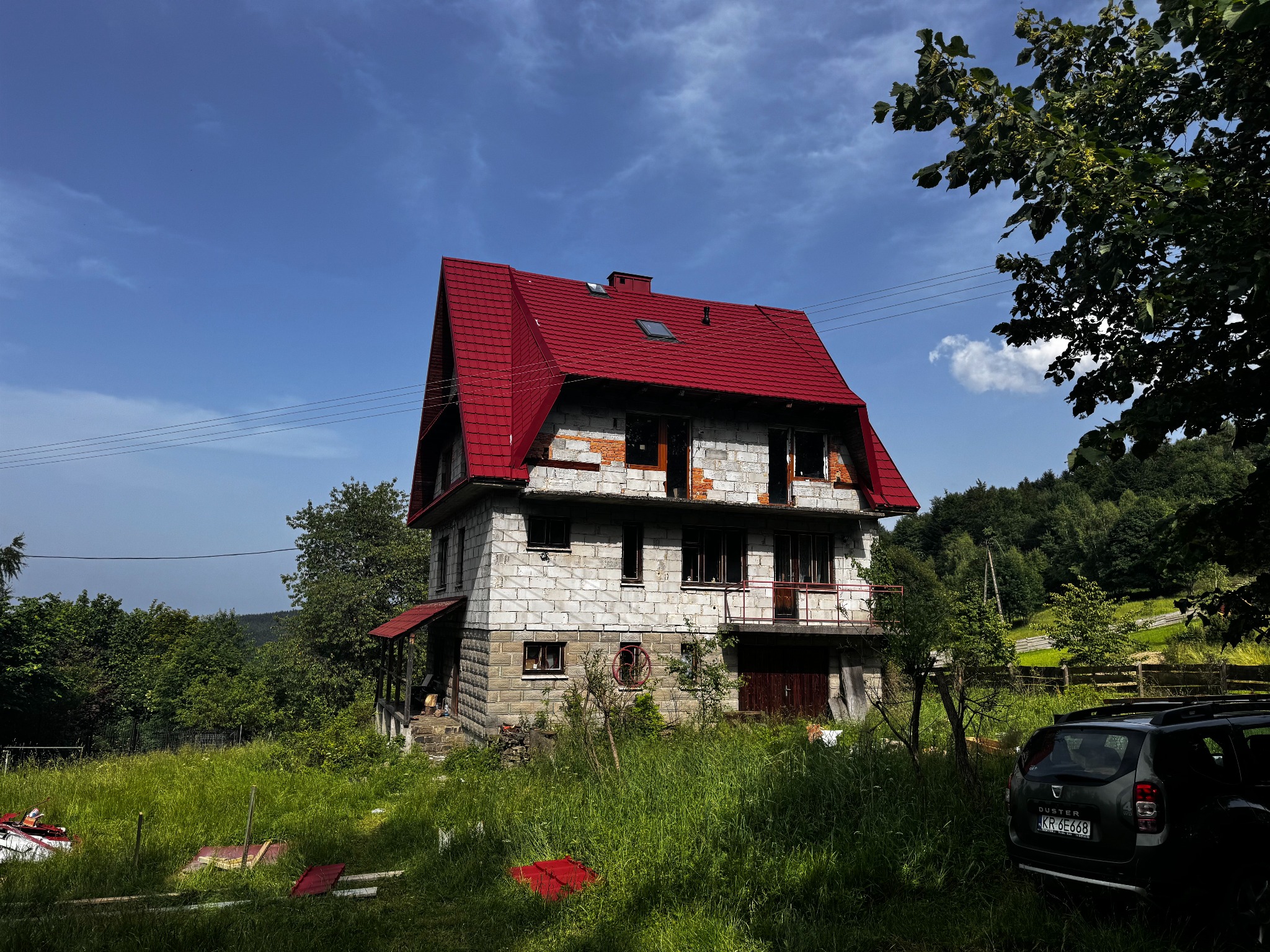 Dwupiętrowy dom w trakcie remontu z intensywnie czerwonym pokryciem dachowym, częściowo otynkowany, otoczony wysoką trawą i drzewami na tle błękitnego nieba.
