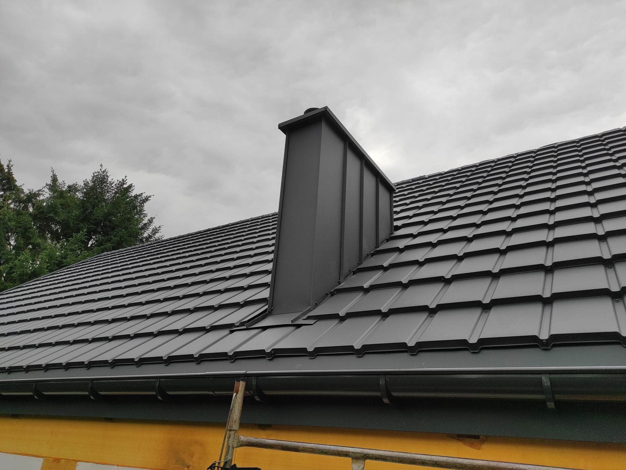 Nowy, ciemnoszary dach z blachodachówki modułowej i obróbka komina z blachy na tle pochmurnego nieba, widoczne rynny i fragment rusztowania.
