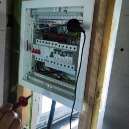 SGP Sp z o.o. - Otwarta skrzynka rozdzielcza z zainstalowanymi bezpiecznikami i przewodami, sprawdzana próbnikiem napięcia w surowym wnętrzu budynku.