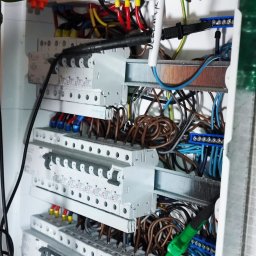 SGP Sp z o.o. - Otwarta skrzynka rozdzielcza z widocznymi bezpiecznikami, przewodami o różnych kolorach i miernikiem w trakcie pomiaru.