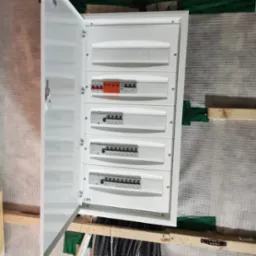 Otwarta biała skrzynka rozdzielcza z widocznymi bezpiecznikami i wyłącznikami, zamontowana na ścianie z izolacją i drewnianymi elementami konstrukcyjnymi.