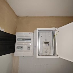 SGP Sp z o.o. - Dwa białe panele elektryczne zamontowane na ścianie, jeden z otwartymi drzwiczkami ukazującymi licznik energii elektrycznej, drugi z częściowo otwartymi drzwiczkami z widocznymi wyłącznikami.