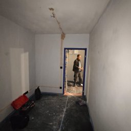 SGP Sp z o.o. - Przygotowanie do prac elektrycznych w Krośnicy: widoczne kable wystające z sufitu, bruzdy na ścianie pod instalację elektryczną, podłoga zabezpieczona folią, elektryk w tle.