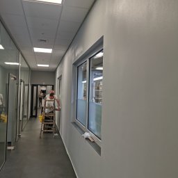 SGP Sp z o.o. - Malowanie ścian w biurowym korytarzu: dwóch mężczyzn pracuje na drabinie, odświeżając szare ściany obok okna z mleczną folią, widoczne żółte wiadro i oświetlenie sufitowe.