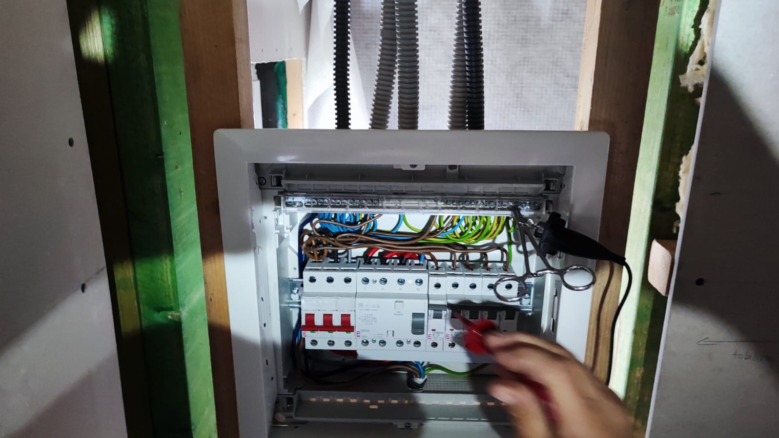 Otwarta skrzynka elektryczna w trakcie pomiarów, widoczne kolorowe przewody, miernik i fragment ręki elektryka z czerwonym próbnikiem, surowe wykończenie wnętrza.