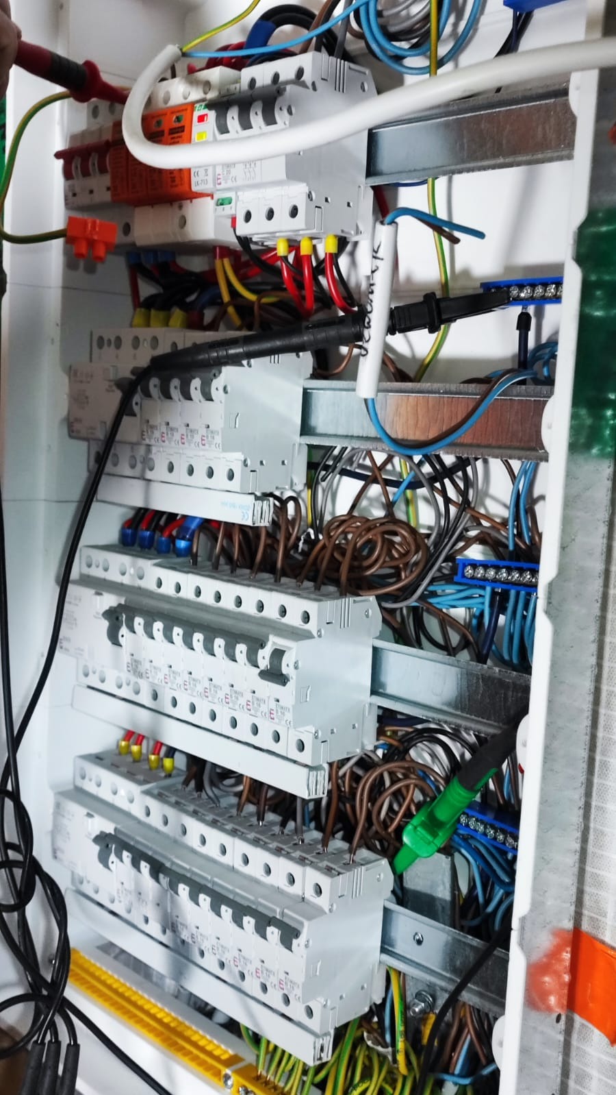 Otwarta skrzynka rozdzielcza z widocznymi bezpiecznikami, przewodami o różnych kolorach i miernikiem w trakcie pomiaru.