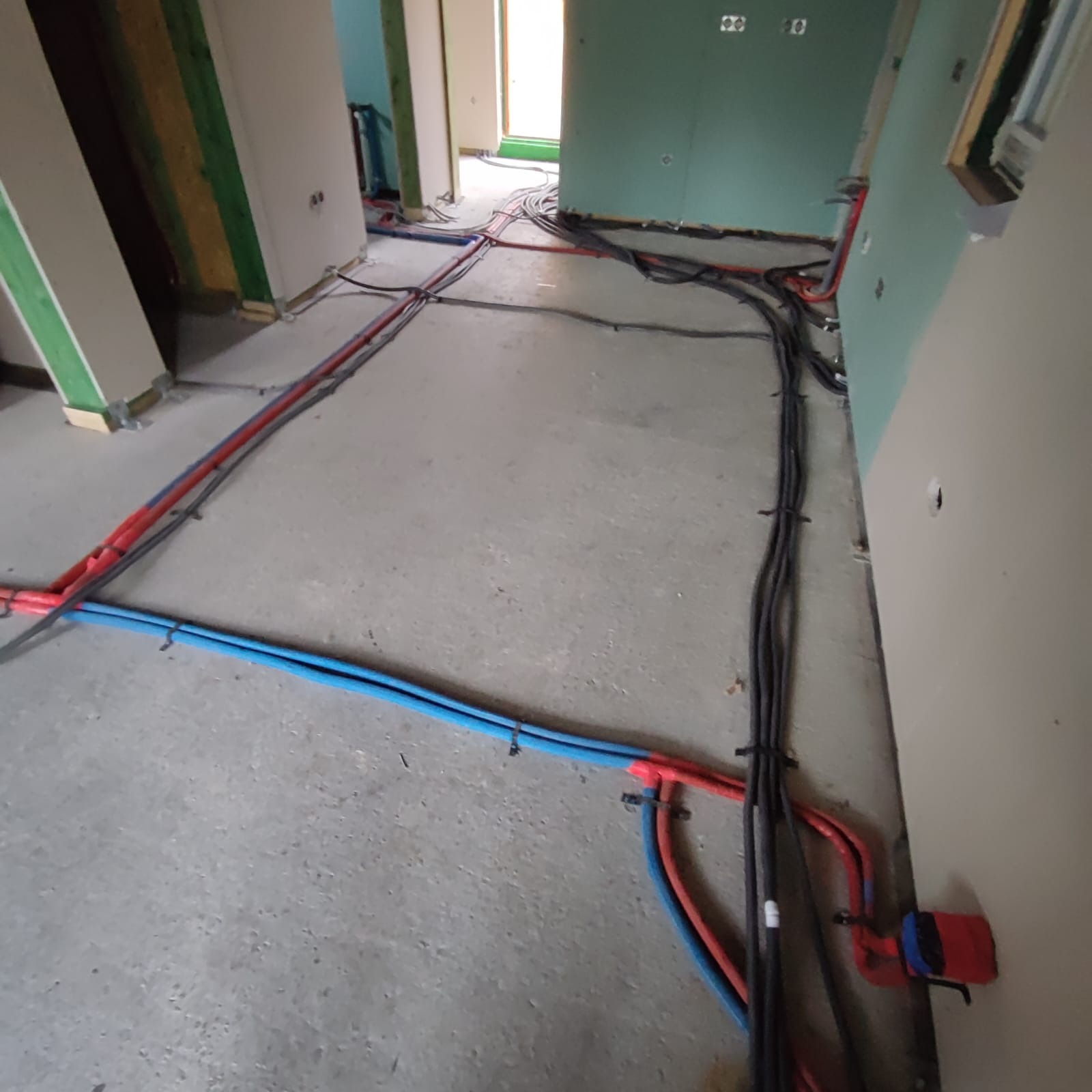 Rozłożone instalacje elektryczne i hydrauliczne w surowym stanie budynku: czarne kable elektryczne i niebiesko-czerwone rury wodne na betonowej podłodze, ściany w stanie deweloperskim z widocznymi...