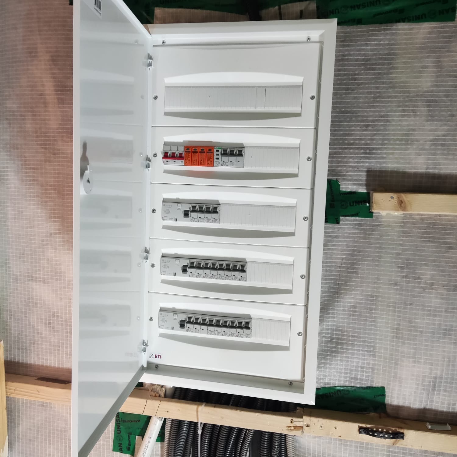 Otwarta biała skrzynka rozdzielcza z widocznymi bezpiecznikami i wyłącznikami, zamontowana na ścianie z izolacją i drewnianymi elementami konstrukcyjnymi.