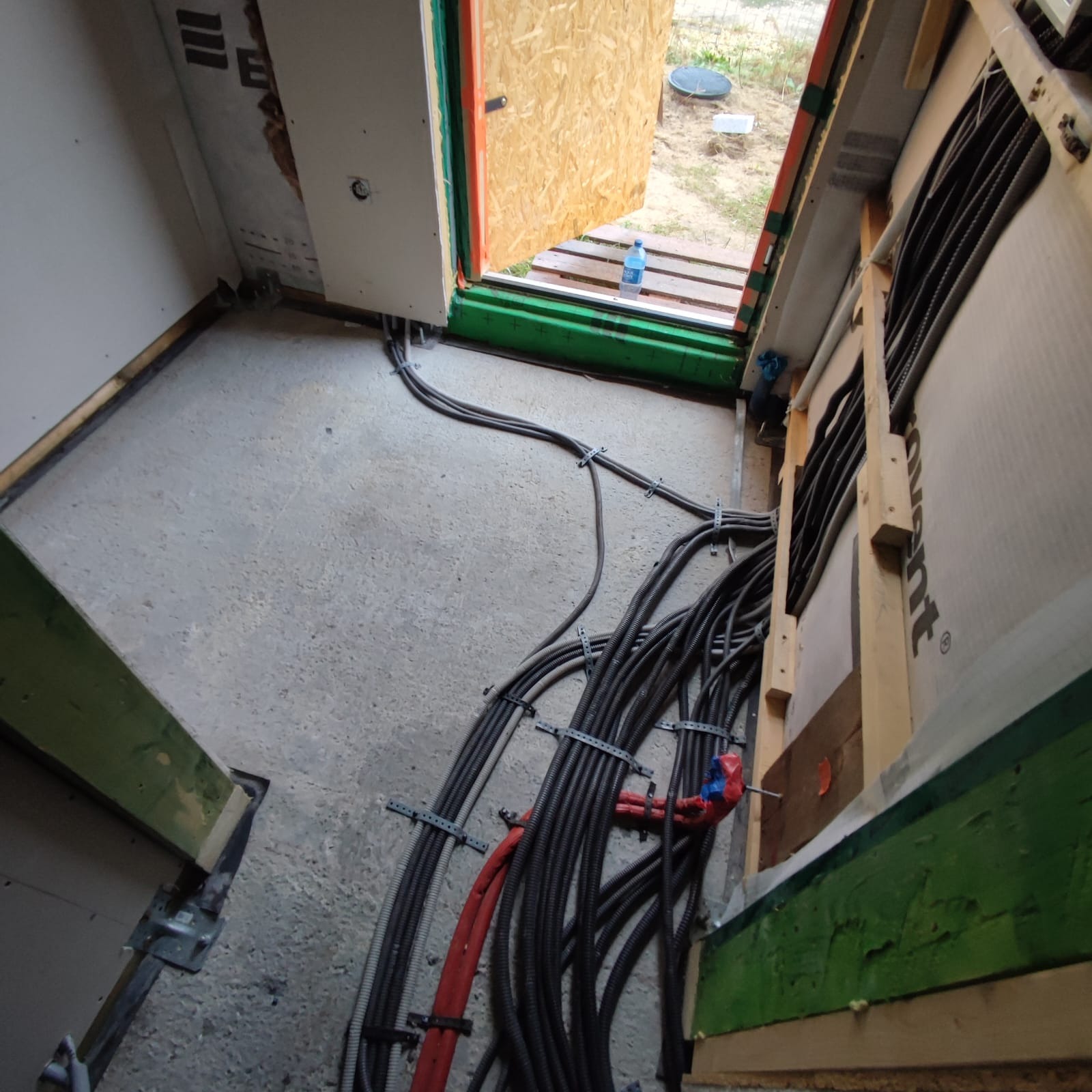 Wiązki kabli elektrycznych przymocowane do ściany w surowym stanie, widoczne przewody w peszlach, mocowania metalowe, fragment izolacji, w tle otwór drzwiowy z butelką wody.