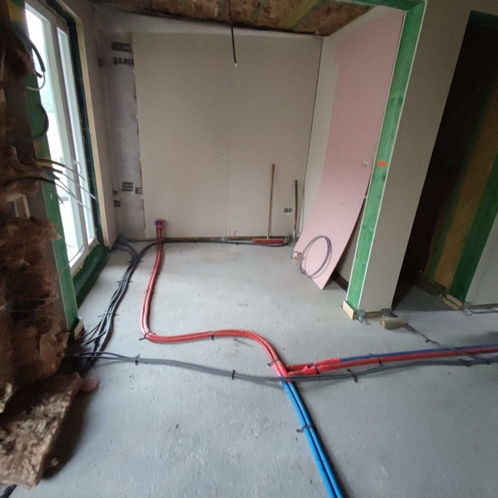 Wewnętrzne instalacje: czerwone i niebieskie rury oraz czarne kable elektryczne prowadzone po podłodze w trakcie remontu w Krośnicy. Widoczne fragmenty ścian z płyt gipsowo-kartonowych i ocieplenia.