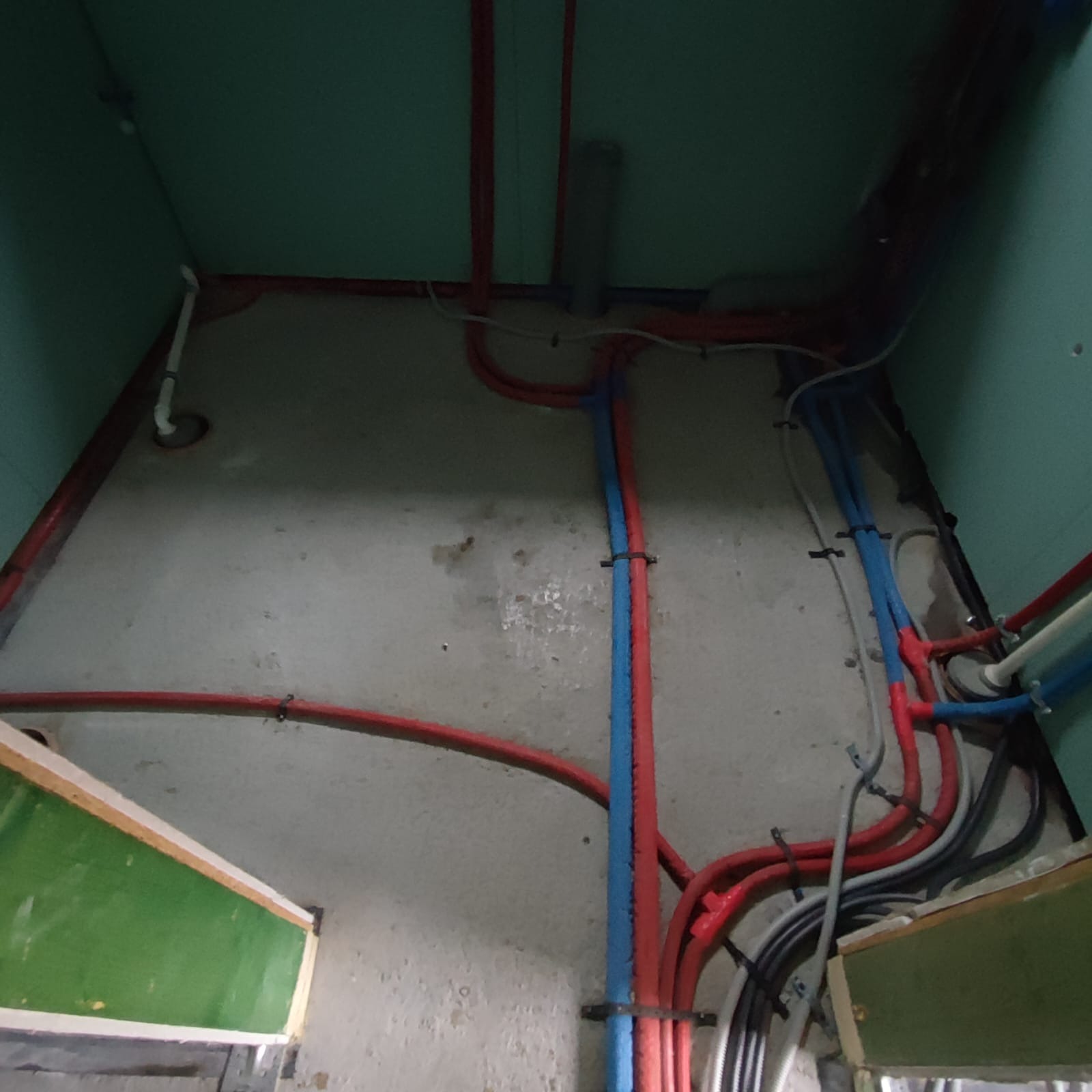 Widok z dołu na ściany i sufit z kolorowymi rurami (czerwone, niebieskie, szare) przymocowanymi do ściany, prawdopodobnie w trakcie instalacji.