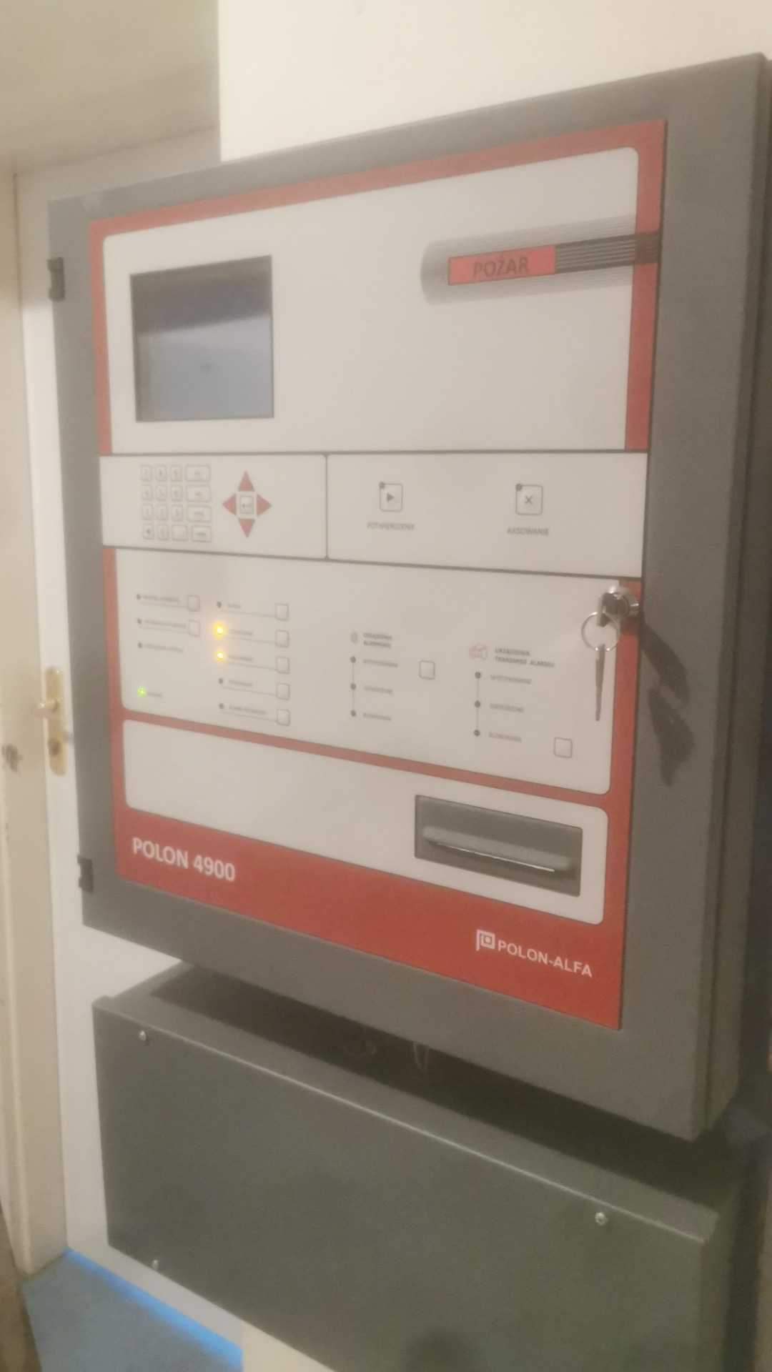 Panel sterowania systemem alarmu pożarowego Polon 4900 z kluczem w zamku, sygnalizujący stan alarmu.