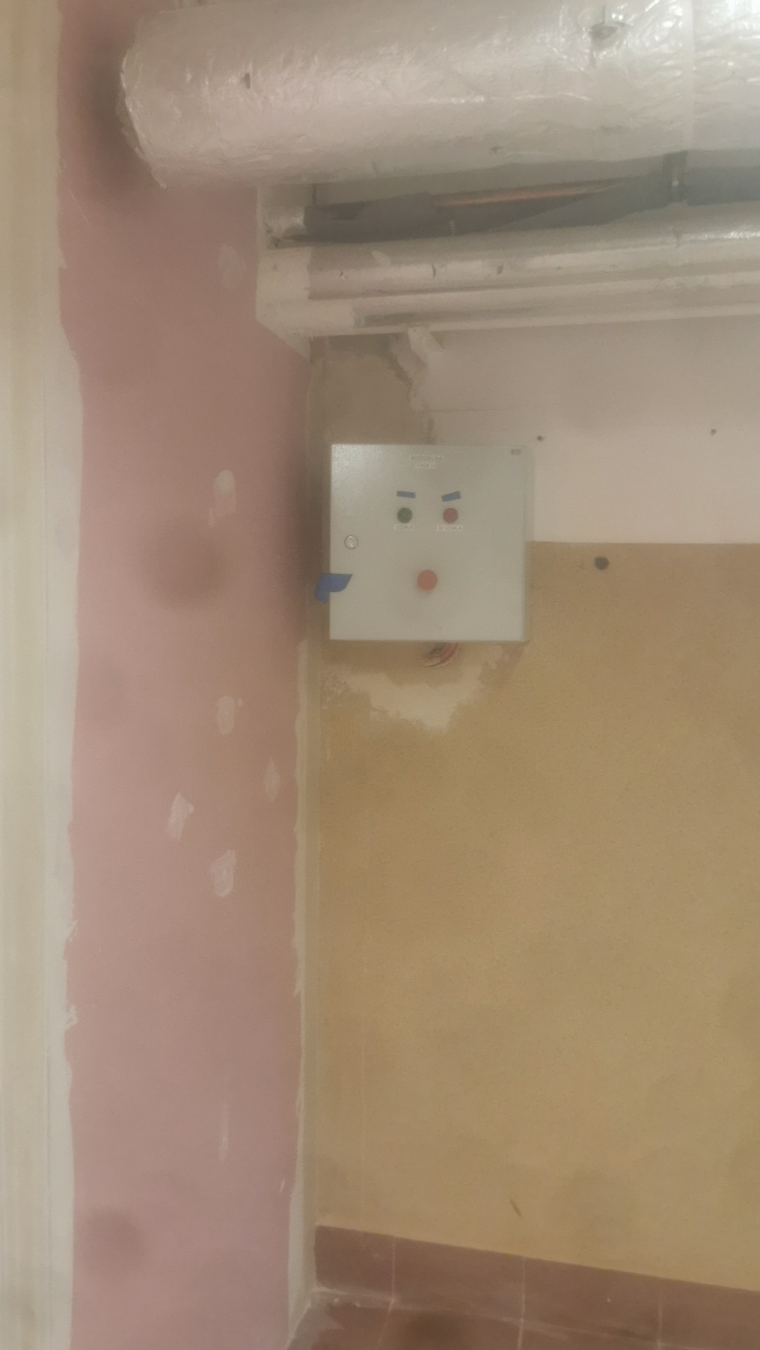 Szara skrzynka elektryczna z trzema okrągłymi elementami (czerwony, zielony, zielony) zamontowana na ścianie w surowym stanie, widoczne rury instalacyjne pod sufitem.