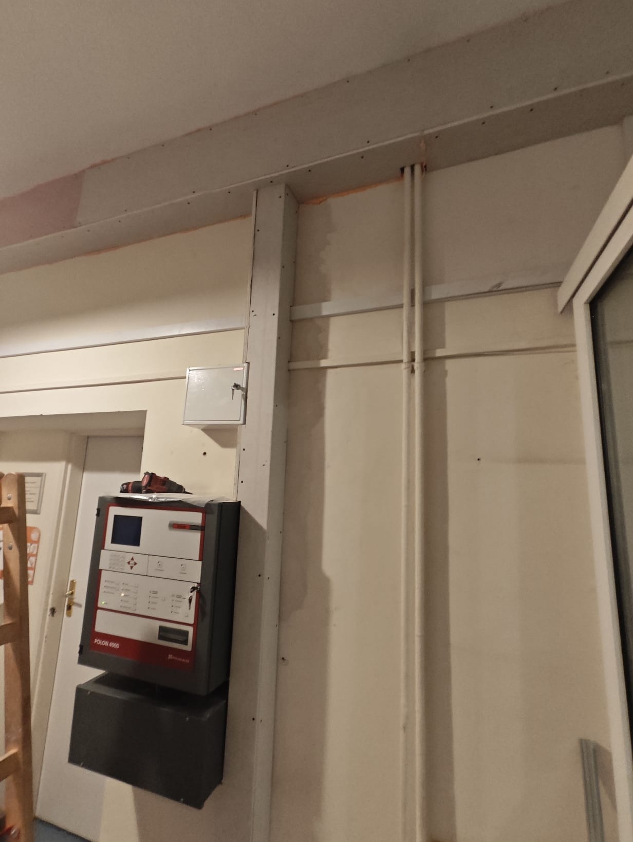 Panel sterowania systemem alarmowym Polon 4900 zamontowany na ścianie, widoczne przewody i narzędzia, w tle drzwi i fragment drabiny.