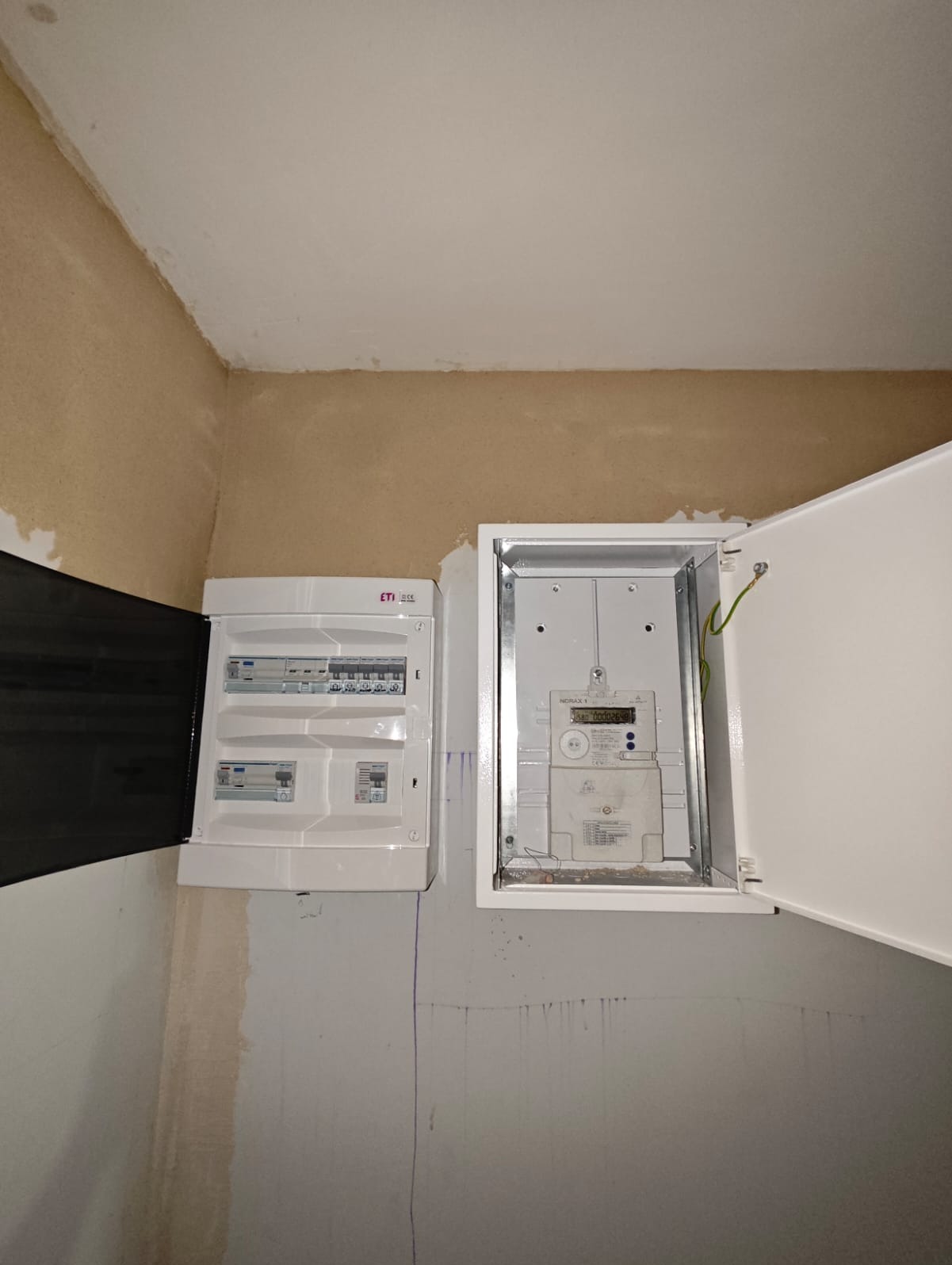 Dwa białe panele elektryczne zamontowane na ścianie, jeden z otwartymi drzwiczkami ukazującymi licznik energii elektrycznej, drugi z częściowo otwartymi drzwiczkami z widocznymi wyłącznikami.