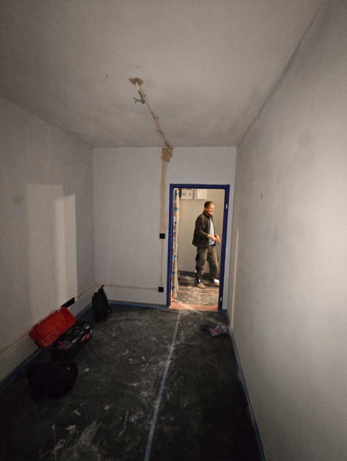 Przygotowanie do prac elektrycznych w Krośnicy: widoczne kable wystające z sufitu, bruzdy na ścianie pod instalację elektryczną, podłoga zabezpieczona folią, elektryk w tle.