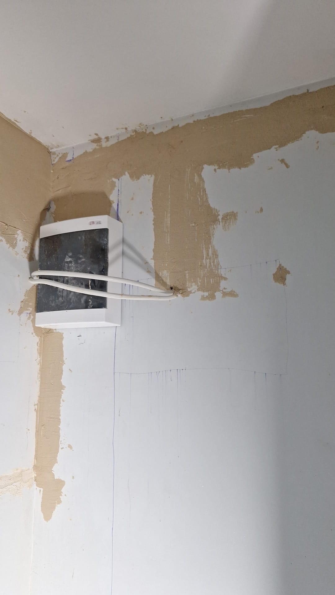 Biała skrzynka elektryczna z przewodami na ścianie w trakcie remontu, widoczne ślady po gładzi i niebieskie linie pomocnicze.