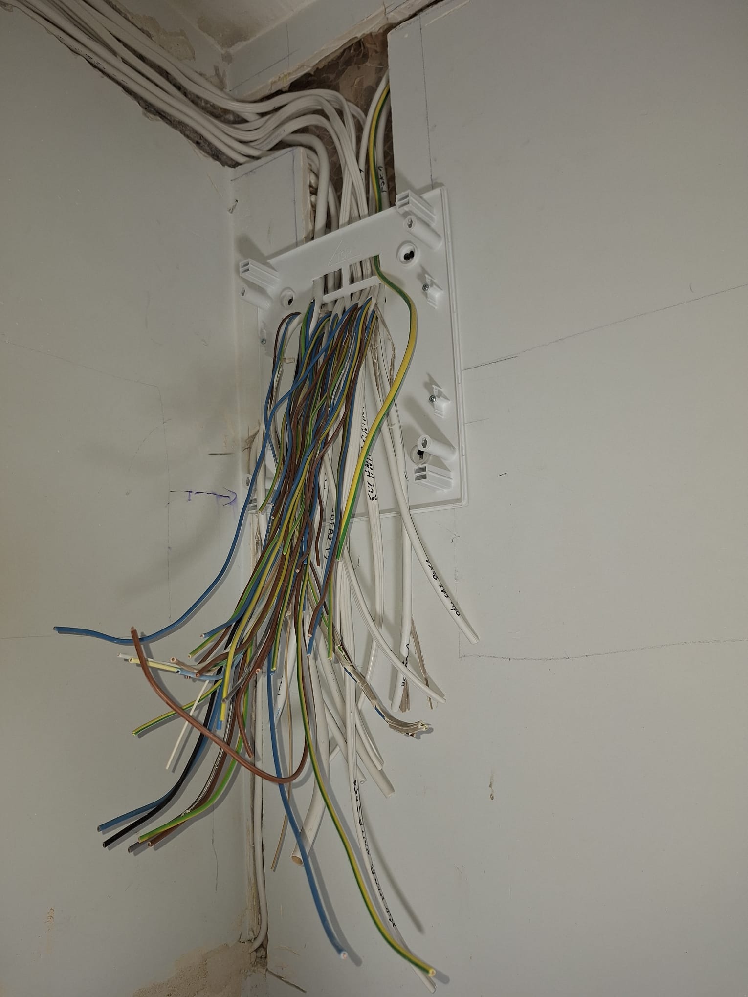 Niezakończona instalacja elektryczna w rogu pomieszczenia, widoczne kable wychodzące z białej puszki montażowej na ścianie, niektóre kable opisane markerem, ściana pomalowana na biało z widocznymi...
