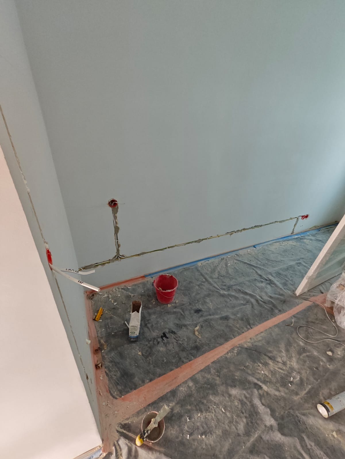 Świeżo położone przewody elektryczne w bruzdach na ścianie, przygotowane do dalszych prac wykończeniowych, zabezpieczona folią podłoga, widoczne narzędzia i wiadro.