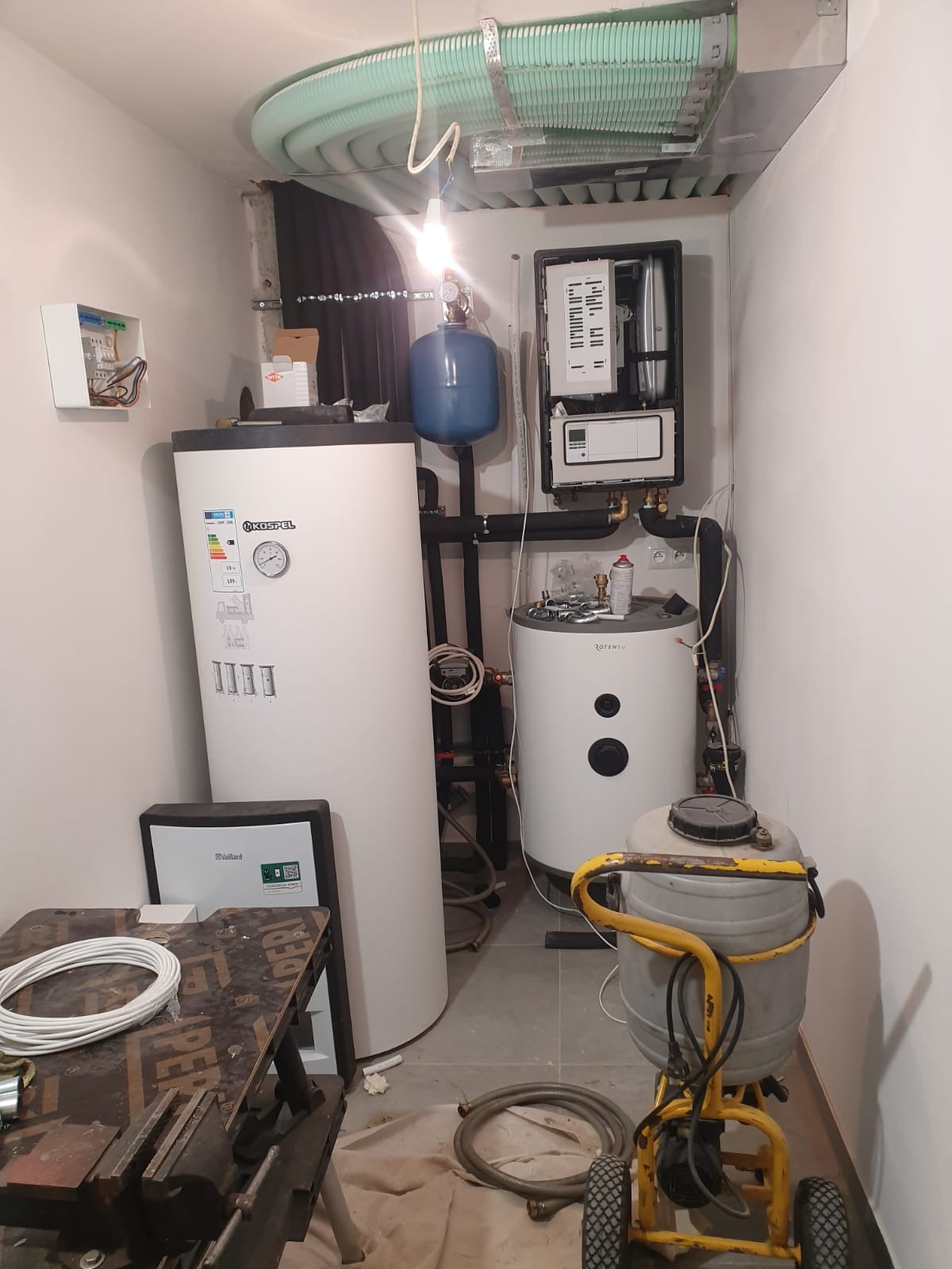 Kompaktowa kotłownia z dwoma białymi zbiornikami, niebieskim naczyniem wzbiorczym, skrzynką elektryczną na ścianie i narzędziami na podłodze, w tym wózkiem z szarym zbiornikiem.