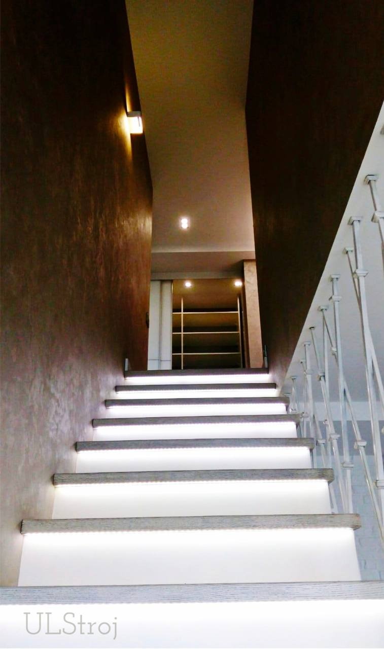 Schody z podświetleniem LED wzdłuż stopni, ściany w odcieniach brązu z dekoracyjną fakturą, biała balustrada oraz widoczna w tle zabudowa meblowa z oświetleniem.