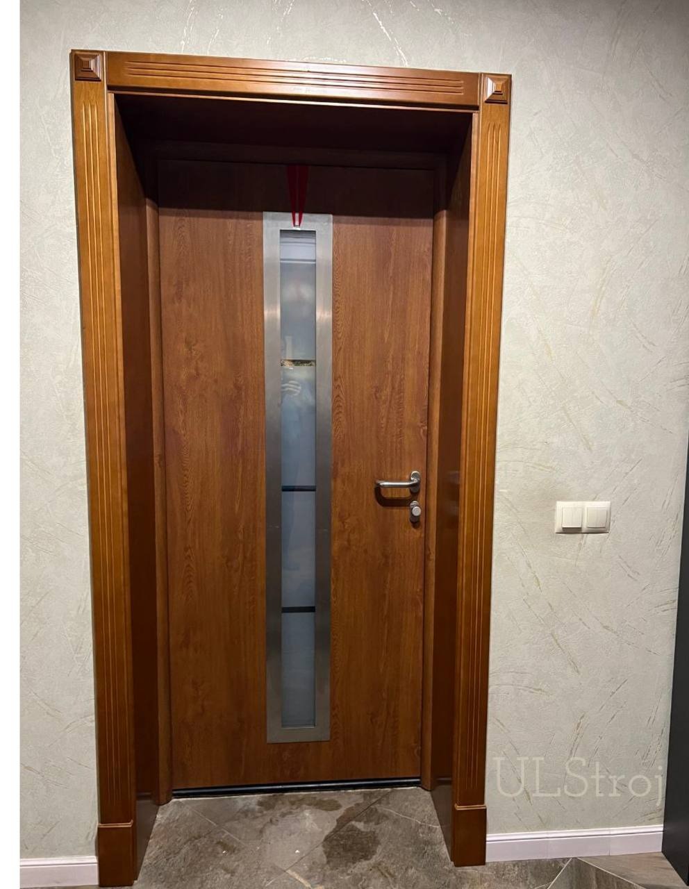 Zamknięte drewniane drzwi wewnętrzne z pionowym, matowym przeszkleniem w metalowej ramie, osadzone w drewnianej ościeżnicy z dekoracyjnymi listwami, na tle ściany z tapetą w jasnym odcieniu...