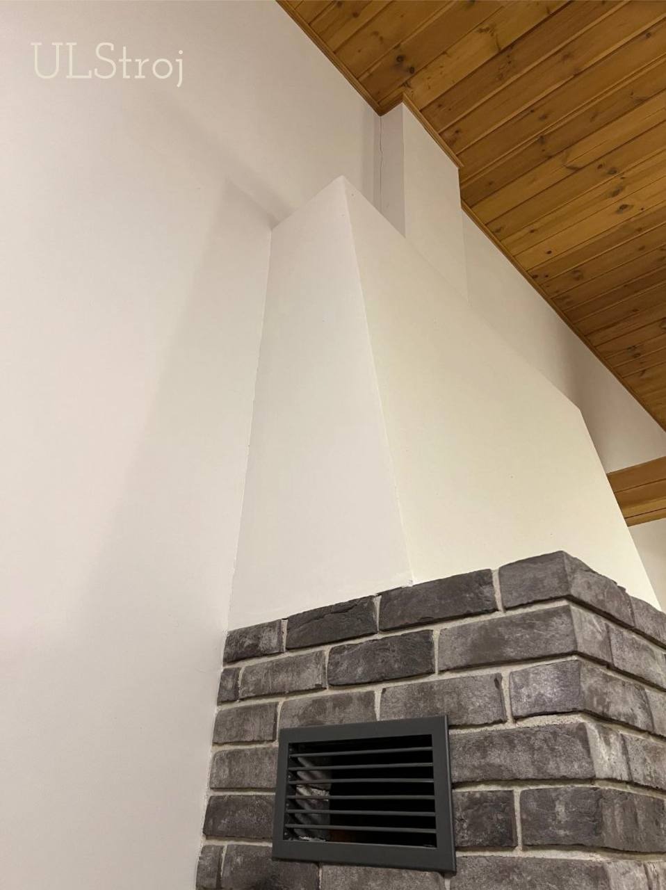 Komin obłożony szarą cegłą klinkierową z kratką wentylacyjną, białe ściany i sufit z drewnianych paneli w tle.