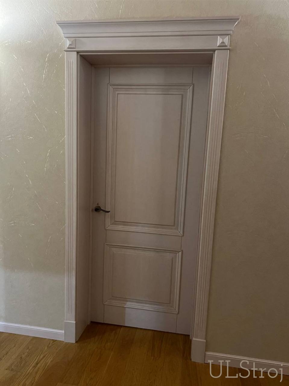 Zamknięte, kremowe drzwi wewnętrzne z dekoracyjną ościeżnicą i listwami przypodłogowymi w jasnym wnętrzu z drewnianą podłogą.