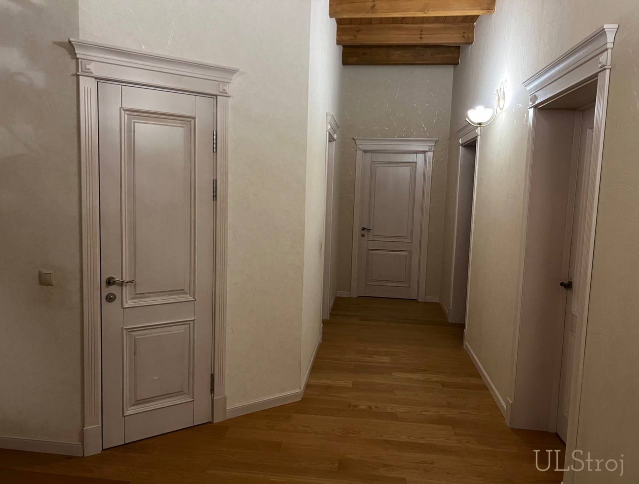 Korytarz z trzema białymi drzwiami z dekoracyjnymi ościeżnicami, drewnianą podłogą i sufitem z drewnianymi belkami.