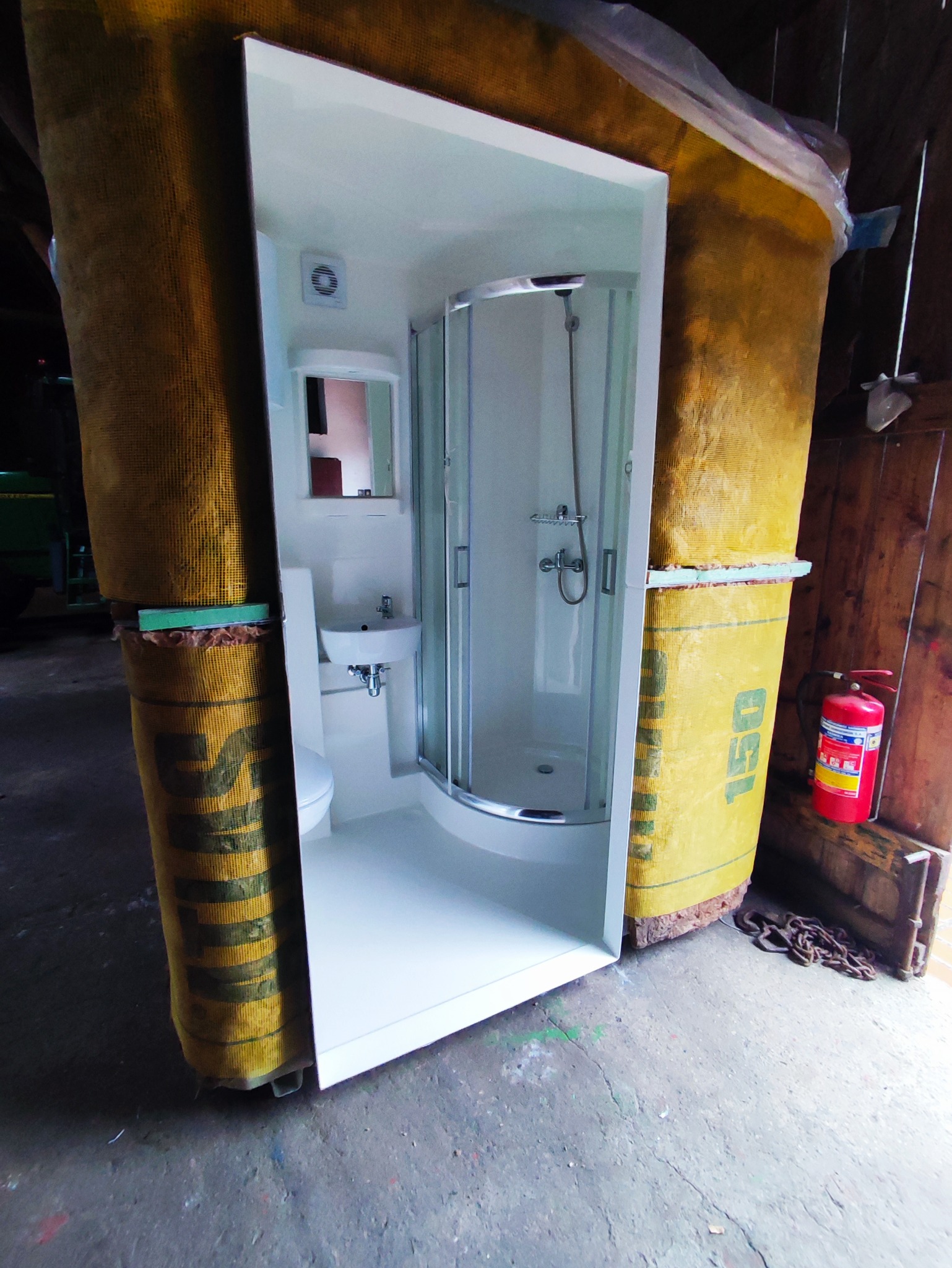 Gotowa kabina prysznicowa z toaletą i umywalką, otoczona materiałem zabezpieczającym, ustawiona w pomieszczeniu gospodarczym z betonową podłogą i gaśnicą w tle.