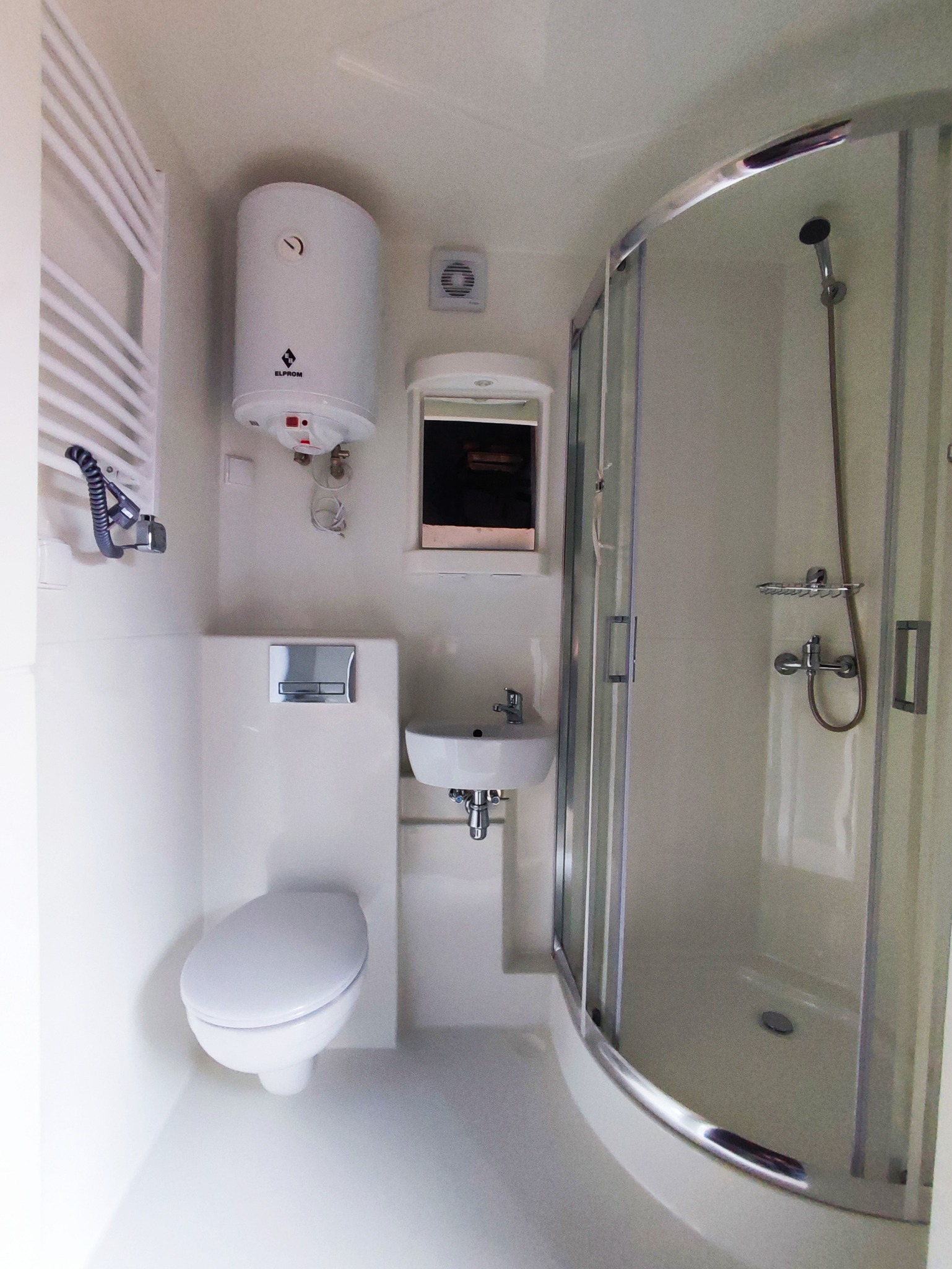 Wnętrze małej, jasnej łazienki z toaletą, umywalką, grzejnikiem drabinkowym, bojlerem i kabiną prysznicową z chromowanymi detalami.