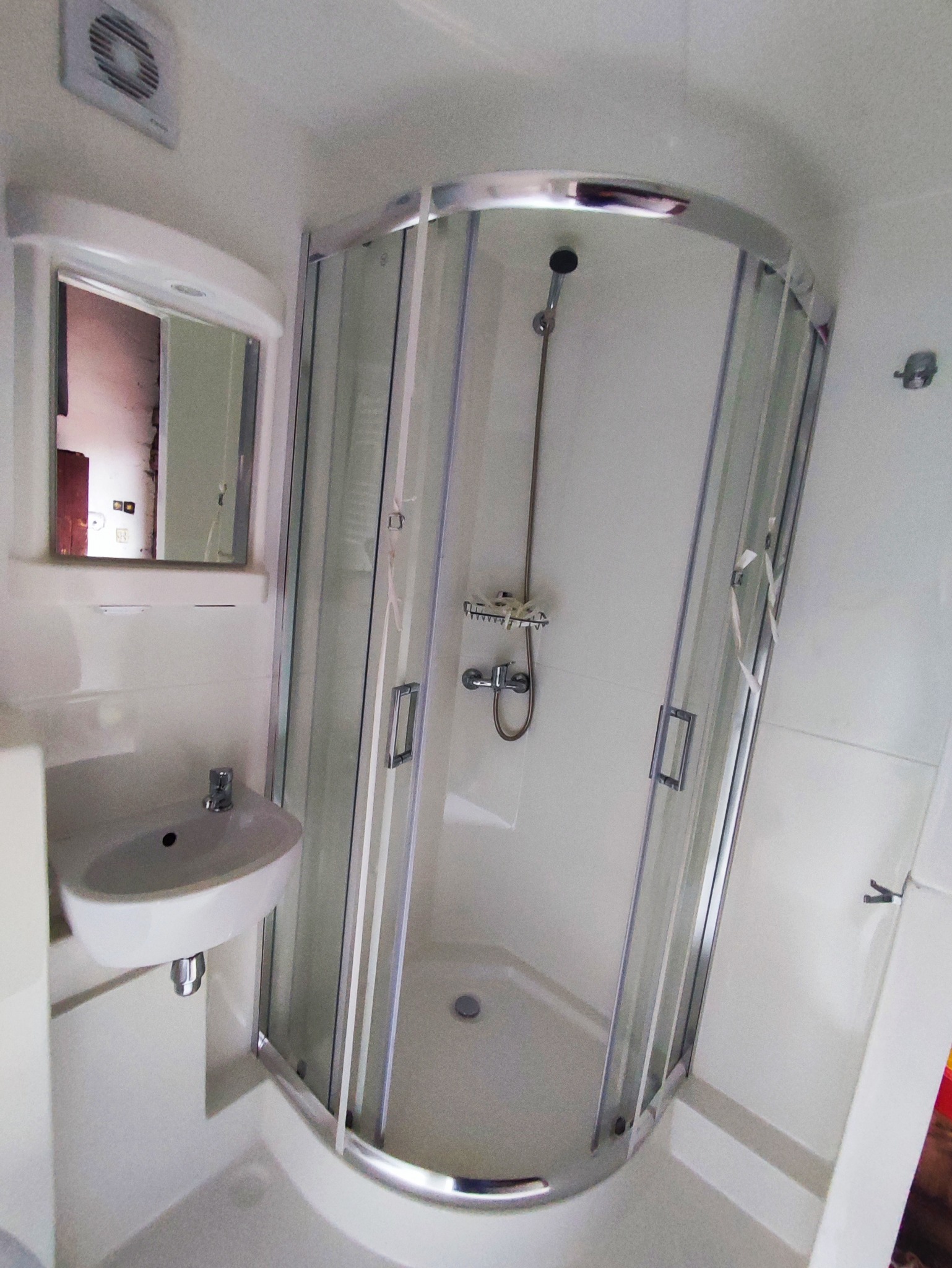 Niewielka, jasna łazienka z narożną kabiną prysznicową, umywalką i lustrem; widoczne ślady po taśmie malarskiej na kabinie.