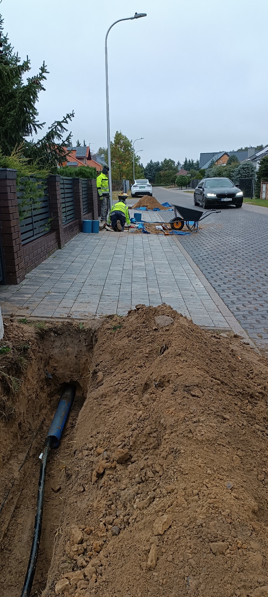 Wykop z widoczną niebieską rurą osłonową i kablami, dwóch pracowników w odblaskowych kamizelkach łączy kable na chodniku z kostki, w tle ulica z samochodami.