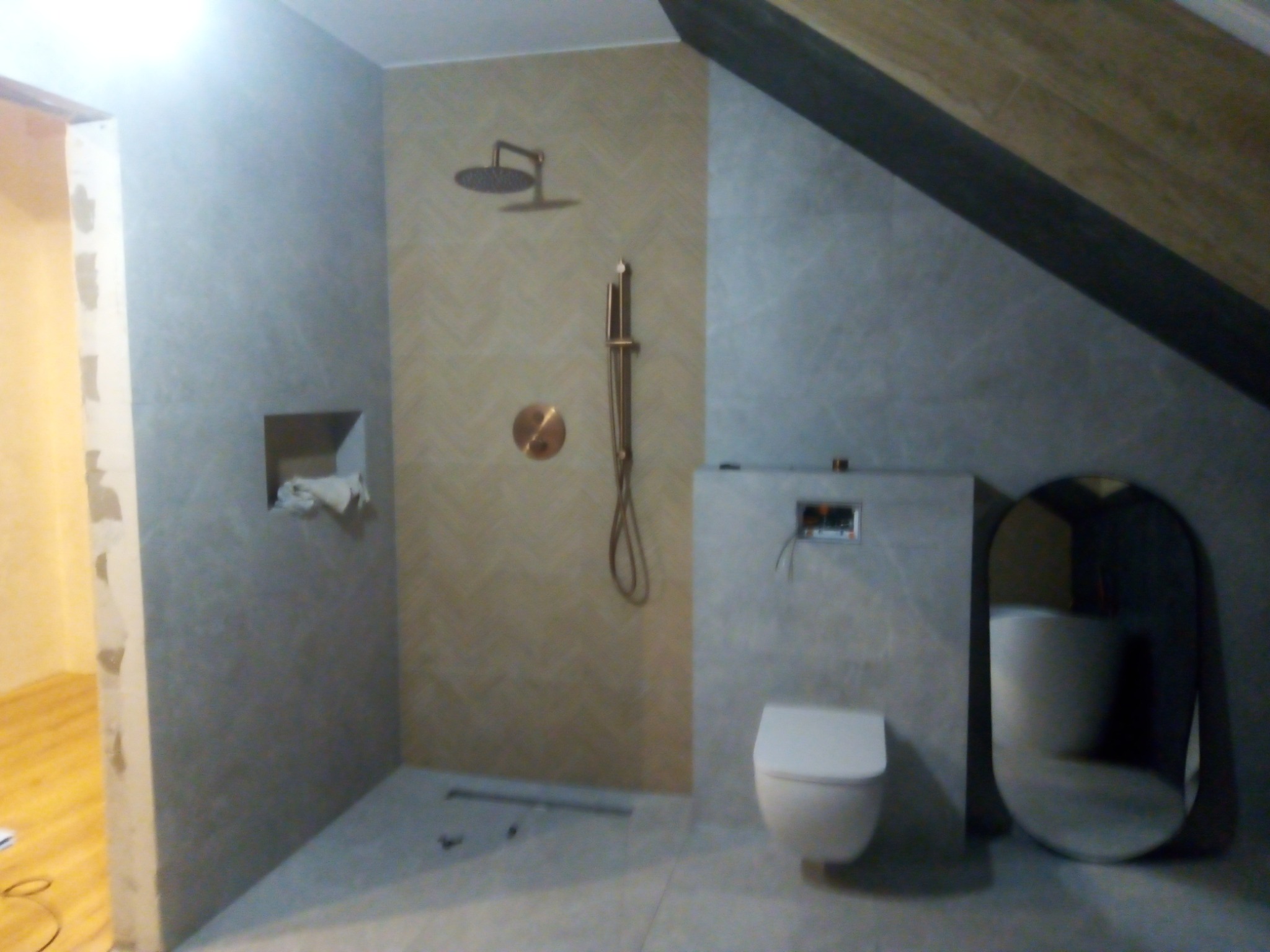 Nowoczesna łazienka w trakcie remontu, z widoczną instalacją prysznicową w kolorze miedzianym na ścianie wyłożonej płytkami w jodełkę, wisząca toaleta i owalne lustro, w tle skośny sufit.