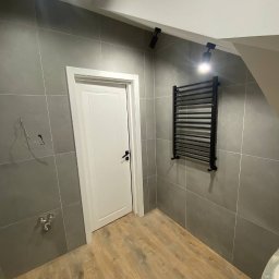 DK Renovations Homes - Wykończona łazienka z szarą, kwadratową glazurą, drewnopodobną podłogą, białymi drzwiami z czarną klamką i czarnym grzejnikiem drabinkowym podświetlonym przez reflektor punktowy.
